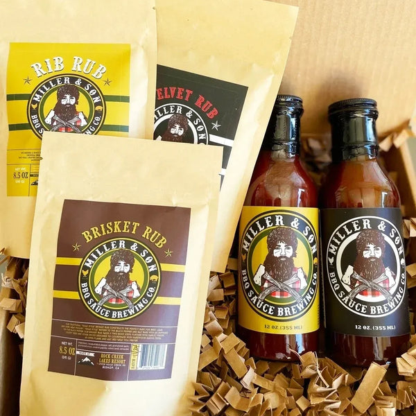 Miller & Son BBQ sauce and rub seasoning products arranged in a gift box with shredded paper filler. The set includes two bottles of sauce with yellow and black labels and three bags of rub seasoning in tan packaging with yellow and dark brown labels.