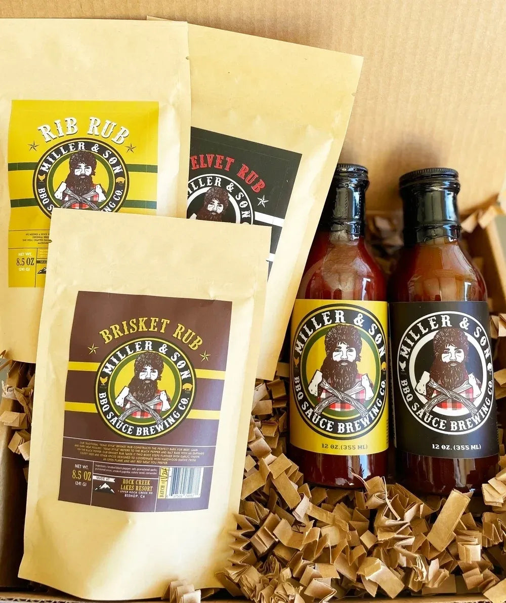 Miller & Son BBQ sauce and rub seasoning products arranged in a gift box with shredded paper filler. The set includes two bottles of sauce with yellow and black labels and three bags of rub seasoning in tan packaging with yellow and dark brown labels.
