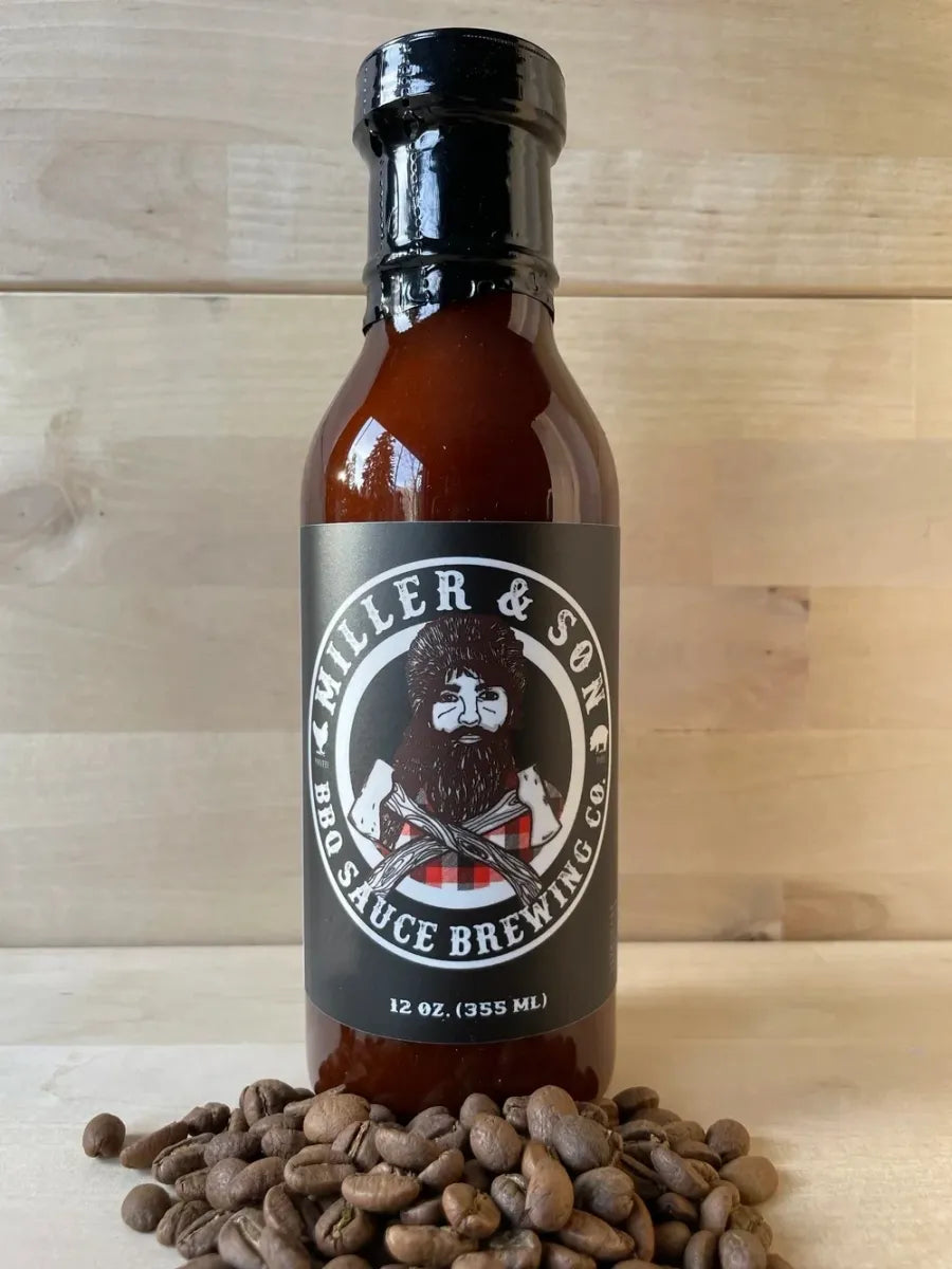 A 12 oz bottle of Miller & Son BBQ Sauce Brewing Co. hot sauce, filled with dark reddish-brown sauce, standing upright on a bed of roasted coffee beans against a light wood plank background.