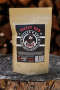 A stand-up pouch of Velvet Rub BBQ Sauce by Miller & Son stands on a rustic tree stump with blurred wood logs in the background. The pouch is a warm tan color with a large black label featuring red and white text and an illustration of a bearded man.