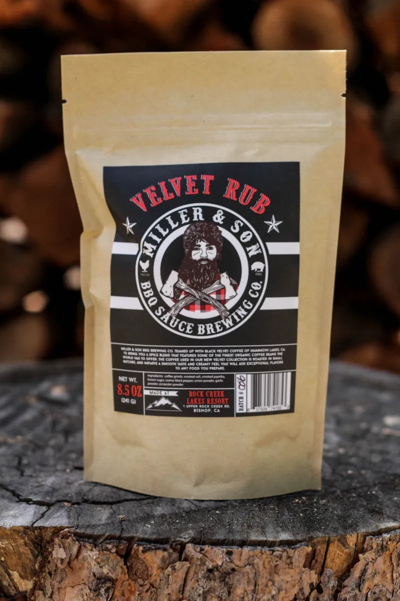 A stand-up pouch of Velvet Rub BBQ Sauce by Miller & Son stands on a rustic tree stump with blurred wood logs in the background. The pouch is a warm tan color with a large black label featuring red and white text and an illustration of a bearded man.