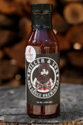A bottle of Killer & Son BBQ Sauce Brewing Co. sauce, 12 oz (355 mL), standing on a rustic wood surface with a blurred background of stacked firewood.