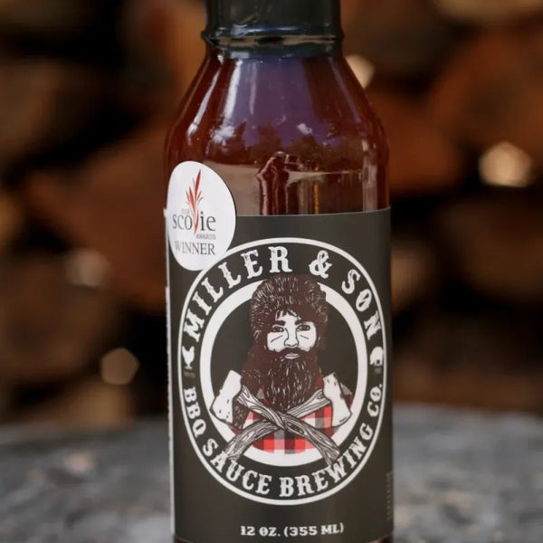 A bottle of Killer & Son BBQ Sauce Brewing Co. sauce, 12 oz (355 mL), standing on a rustic wood surface with a blurred background of stacked firewood.