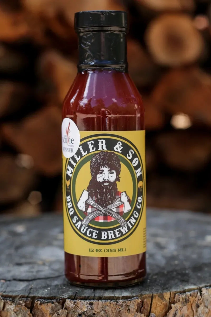 A bottle of Miller & Son BBQ Sauce Brewing Co. hot sauce, filled with reddish-brown sauce, featuring a yellow label with a cartoon bearded man, set on a rustic wood log with stacked firewood in the background.