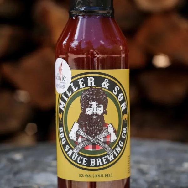 A bottle of Miller & Son BBQ Sauce Brewing Co. hot sauce, filled with reddish-brown sauce, featuring a yellow label with a cartoon bearded man, set on a rustic wood log with stacked firewood in the background.