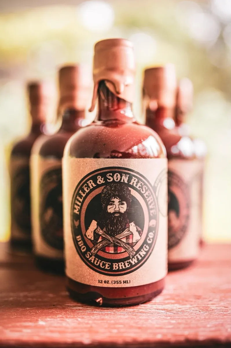 A bottle of Miller & Son Reserve BBQ Sauce Brewing Co. sits on a rustic wooden surface with several matching bottles blurred in the background.