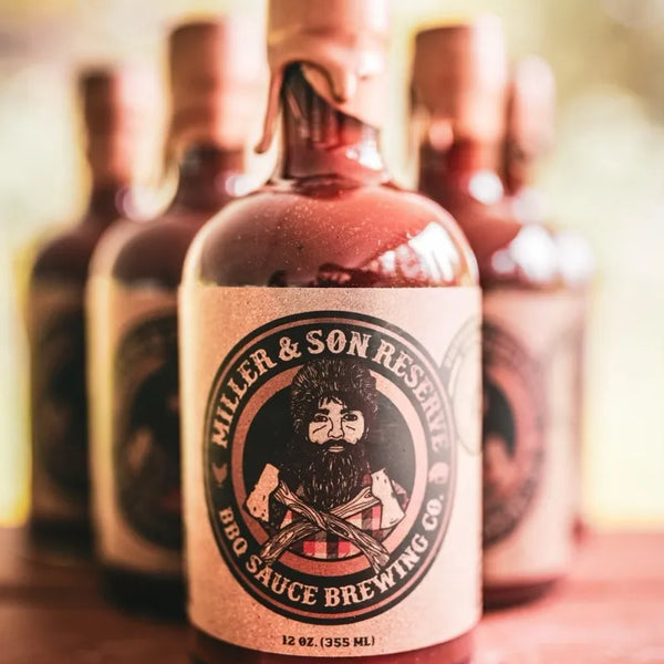 A bottle of Miller & Son Reserve BBQ Sauce Brewing Co. sits on a rustic wooden surface with several matching bottles blurred in the background.