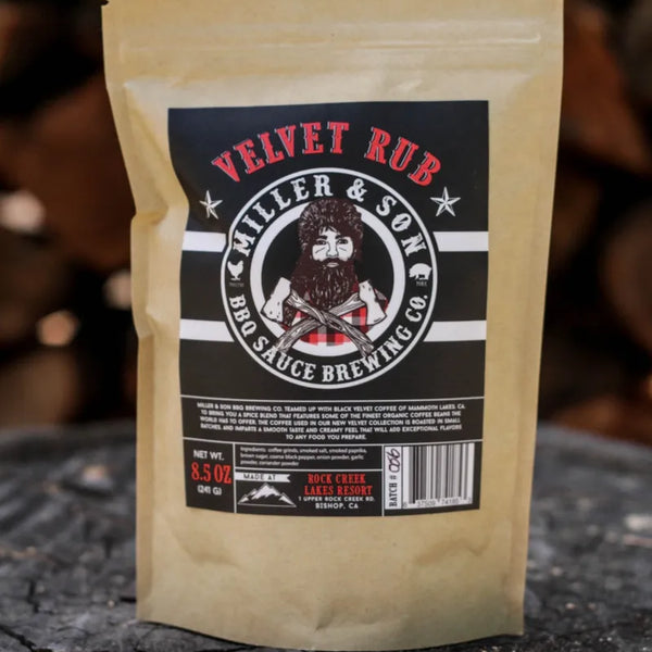 A stand-up pouch of Velvet Rub BBQ Sauce by Miller & Son stands on a rustic tree stump with blurred wood logs in the background. The pouch is a warm tan color with a large black label featuring red and white text and an illustration of a bearded man.
