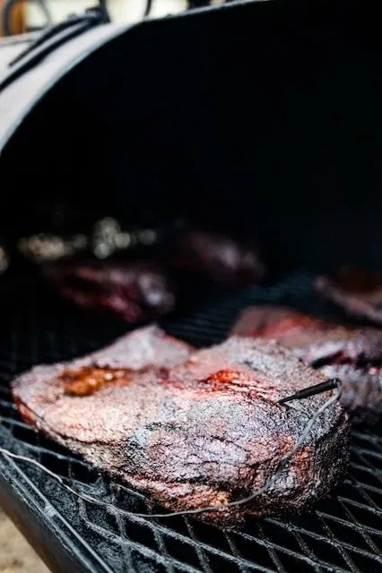 A close-up shot of a piece of smoked meat cooking on a black grill grate, with the lid arching over the meat in the background.