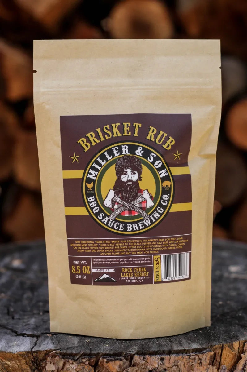 A stand-up kraft paper pouch of Miller & Son Brisket Rub BBQ Sauce Brewing Co. stands on a rustic wood surface with blurred stacked firewood in the background.