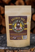 A stand-up kraft paper pouch of Miller & Son Brisket Rub BBQ Sauce Brewing Co. stands on a rustic wood surface with blurred stacked firewood in the background.