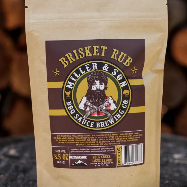 A stand-up kraft paper pouch of Miller & Son Brisket Rub BBQ Sauce Brewing Co. stands on a rustic wood surface with blurred stacked firewood in the background.