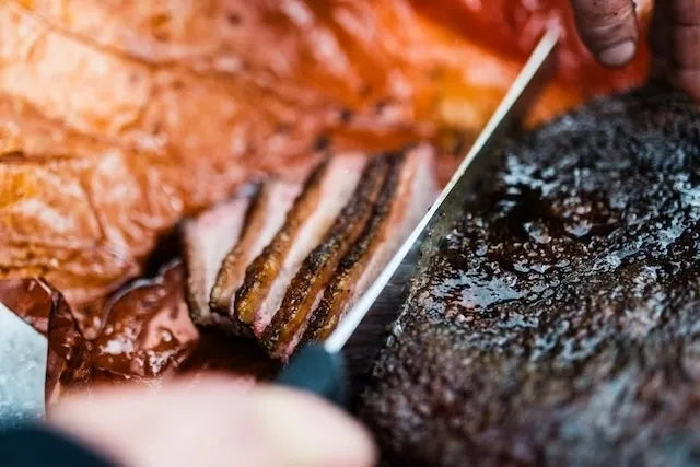 Close-up of sliced smoked meat with a knife, showing pinked interior and dark seared exterior on a rustic surface.