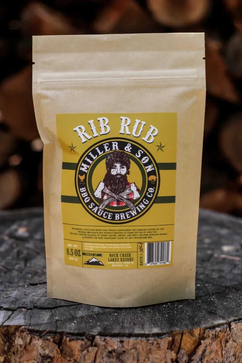 A stand-up kraft paper pouch labeled 'RIB RUB' by Miller & Son BBQ Sauce Brewing Co., placed on a rustic tree stump with blurred wood logs in the background.