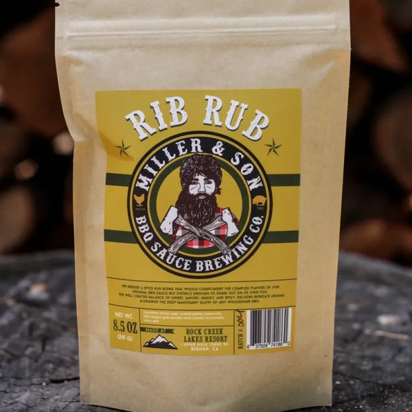 A stand-up kraft paper pouch labeled 'RIB RUB' by Miller & Son BBQ Sauce Brewing Co., placed on a rustic tree stump with blurred wood logs in the background.