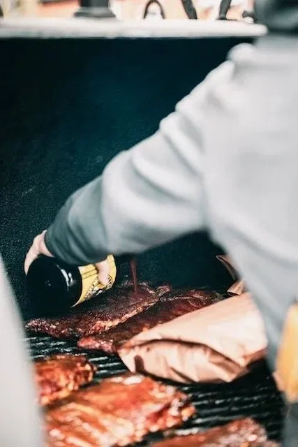 A close-up, over-the-shoulder view of a person pouring dark barbecue sauce from a bottle onto ribs cooking on a grill, with a paper bag of meat beside the ribs.