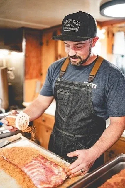 A man in a kitchen wearing a black trucker hat that says 'Support Your Local Smuggler' and a dark denim apron over a gray t-shirt, seasoning raw ribs with a spice shaker.