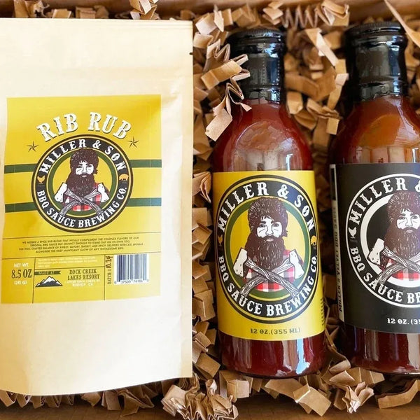 Miller & Son BBQ Sauce Brewing Co. gift set in a box with crinkle paper filler, featuring a white pouch and two bottles of sauce with black and yellow labels.