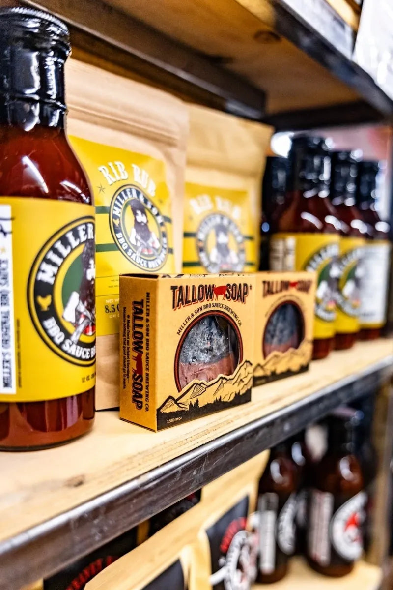 A wooden shelf displays Hillfired Smokehouse hot sauce bottles and Tallow Soap boxes. The products feature bright yellow labels and packaging with black and white branding, set against a dark background with more products blurred.