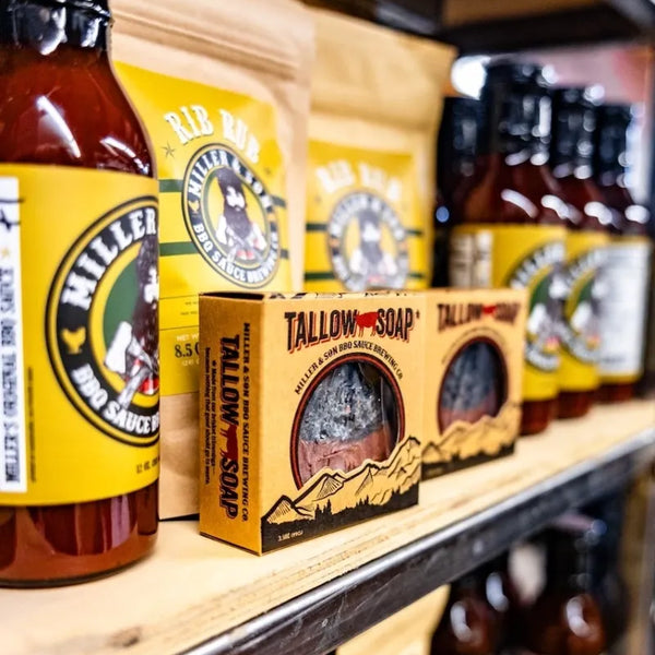 A wooden shelf displays Hillfired Smokehouse hot sauce bottles and Tallow Soap boxes. The products feature bright yellow labels and packaging with black and white branding, set against a dark background with more products blurred.